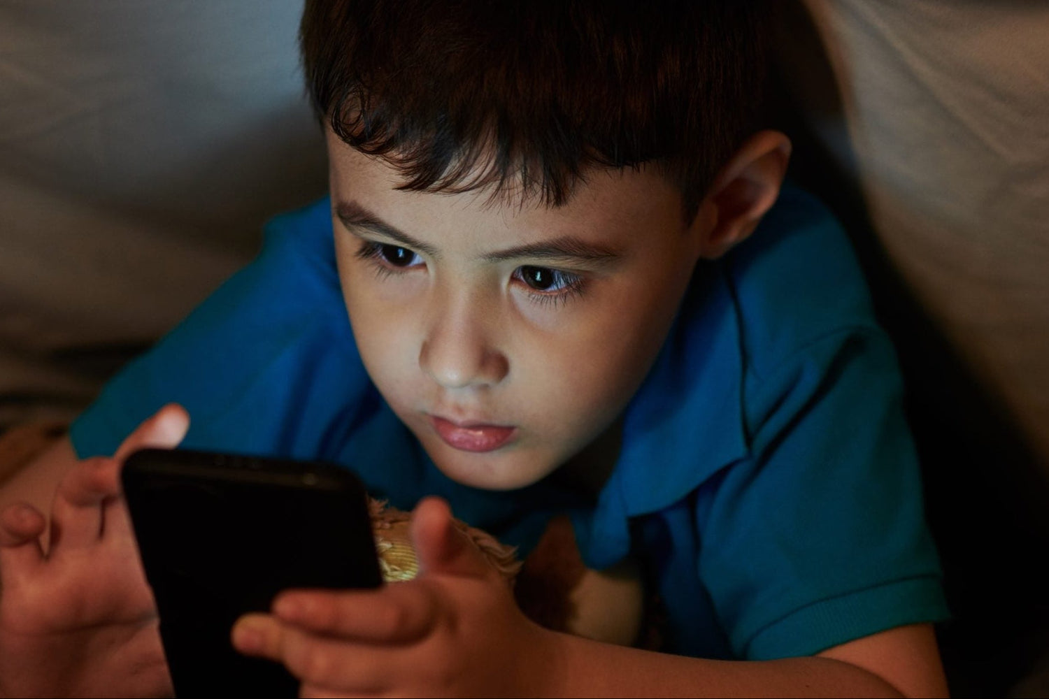 Child in a blue shirt lying on a bed, looking at a smartphone under a blanket.